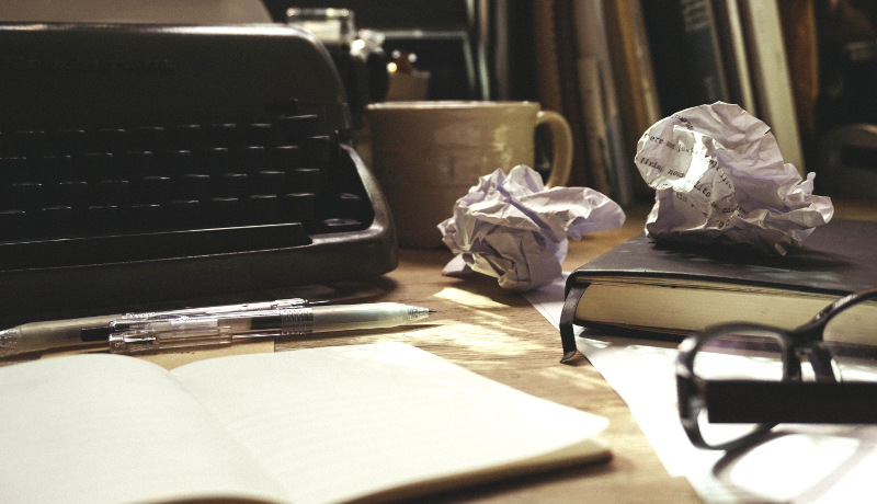 and old typewriter, a notebook and crumpled pages of writing on an aged wooden desk